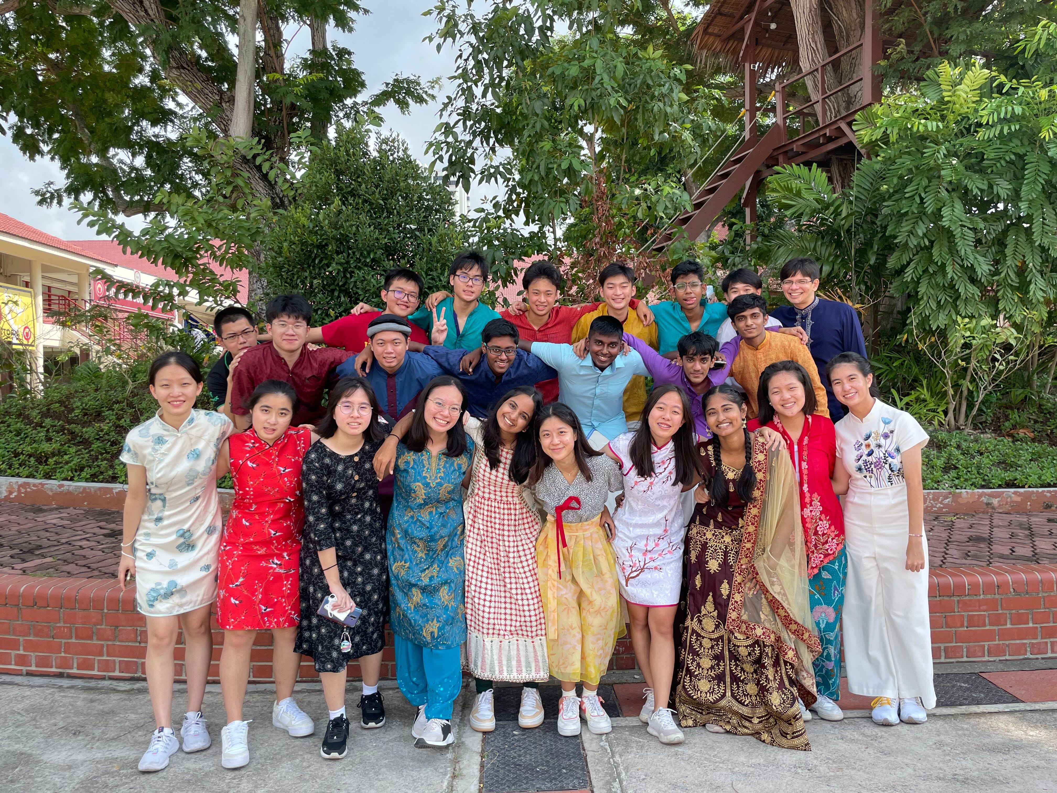 A large group of young people in colorful traditional Asian attire pose together outdoors near trees and a brick wall.